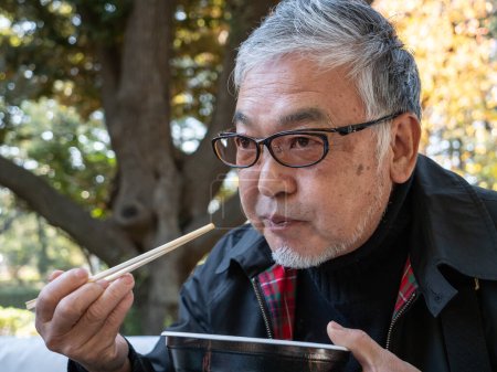 Photo for An older Japanese man eating warm noodles and soup with chopsticks outside - Royalty Free Image