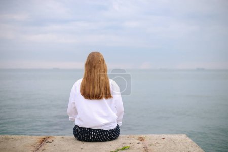 Téléchargez les photos : Vue arrière de belle jeune femme assise sur la jetée et regardant la mer - en image libre de droit