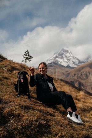Téléchargez les photos : Jeune femme assise sur la colline avec son chien sur le fond des montagnes enneigées - en image libre de droit