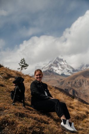 Téléchargez les photos : Fille souriant avec un chien noir dans les montagnes Kazbek en Géorgie en automne. Aventure de voyage - en image libre de droit