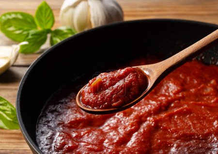 Téléchargez les photos : Ramassez la sauce tomate dans la casserole avec une cuillère. Scène de cuisine. Gros plan. Fond en bois. Basilic et ail en arrière-plan. - en image libre de droit