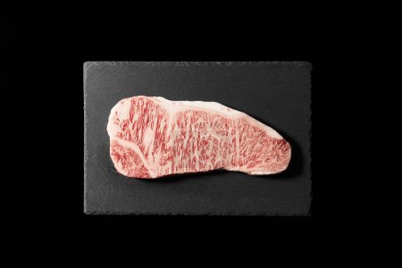Fresh raw beef steak placed on a stone plate against a black background. Wagyu beef steak. View from directly above.