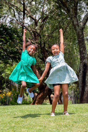 Foto de Full-length portrait of two African girls jumping together in a green garden. Two youngsters holding hands shouting. - Imagen libre de derechos