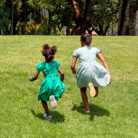 Full-length action portrait of two young African girls having a race in an urban garden. Rear view of kids in green dresses.