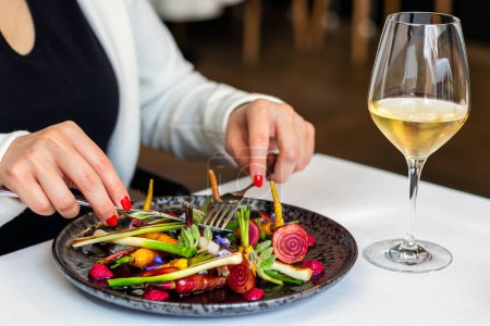 Foto de Detalle de cerca de la mujer comiendo apetitosa ensalada de verduras frescas mezcladas en el restaurante gourmet. Copa de vino blanco en la mesa al lado del plato. - Imagen libre de derechos