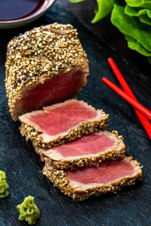 Gros plan vertical de steak de thon croûté au sésame appétissant. Tranches de thon à nageoires jaunes moyennes rares sur ardoise noire avec wasabi, sauce soja et baguettes.