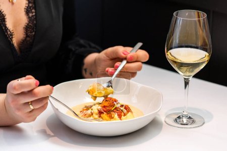 Foto de Detalle de cerca de la mujer cenando en un restaurante gourmet. Manos femeninas sosteniendo cuchara con sopa de langosta junto a un vaso de vino blanco. - Imagen libre de derechos