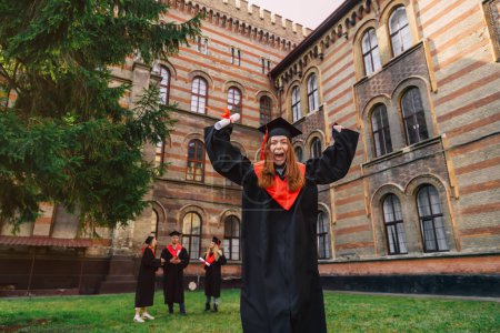 Foto de Bastante sonriente chica graduada universitaria en manto y sombrero saltando con diploma delante de la universidad, compañeros de clase felices celebrando en el fondo. - Imagen libre de derechos