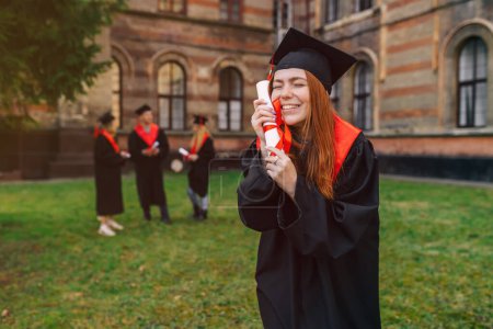 Foto de Chica sonriente, alegre, pelirroja, abrazando un diploma en sus manos. Estudiante en bata de graduación y gorra con pico. - Imagen libre de derechos