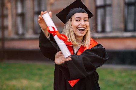 Foto de Una estudiante, de pie sola en el contexto de la universidad, muestra felizmente su diploma, celebra su final de sus estudios. - Imagen libre de derechos