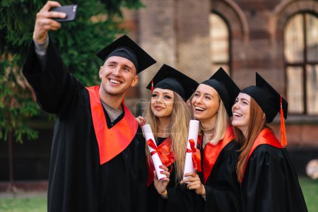 Foto de Concepto de educación, graduación, tecnología y personas - grupo de estudiantes felices en tablas de mortero y vestidos de soltero con diplomas tomando selfie al aire libre. - Imagen libre de derechos