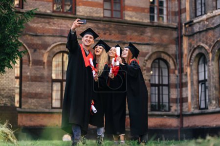 Foto de Grupo de estudiantes felices en batas de soltero con diplomas tomando una selfie en el teléfono al aire libre. - Imagen libre de derechos