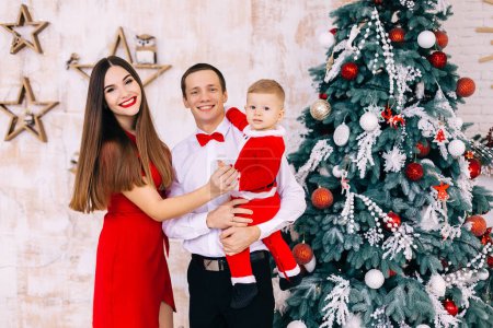 Photo for Family with baby in santa suit look at camera and smile near christmas tree. - Royalty Free Image