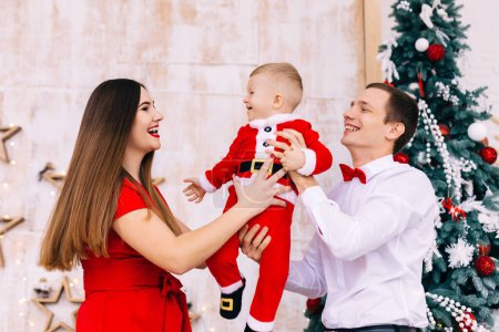 Photo for Dad and mom holding son in their arms in festive clothes near the Christmas tree. - Royalty Free Image