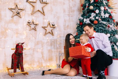 Photo for Baby in santa costume holds big gift box and smile. parents sitting near Christmas tree and smiling. rocking horse in the room. - Royalty Free Image