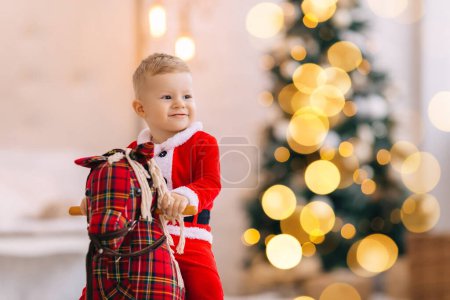 Téléchargez les photos : Le petit garçon est assis sur un cheval à bascule et sourit. arbre de Noël flou en arrière-plan. - en image libre de droit