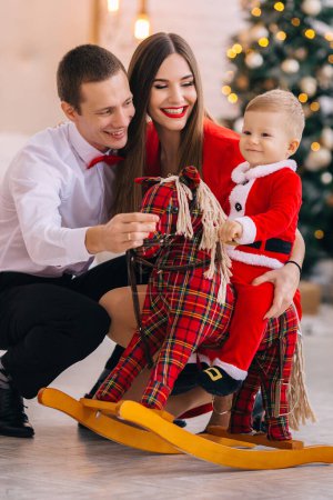 Foto de Bebé balanceándose en el caballo mecedora y los padres sentados y sonriendo. Juega. árbol de Navidad. - Imagen libre de derechos