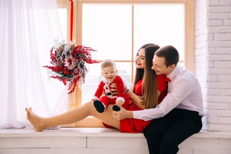 Photo for Parents sitting on windowsill and holding baby. the window is decorated with a Christmas wreath. - Royalty Free Image