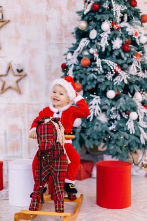Téléchargez les photos : Bébé garçon en costume de Père Noël sur cheval à bascule près de l'arbre de Noël et des cadeaux. - en image libre de droit
