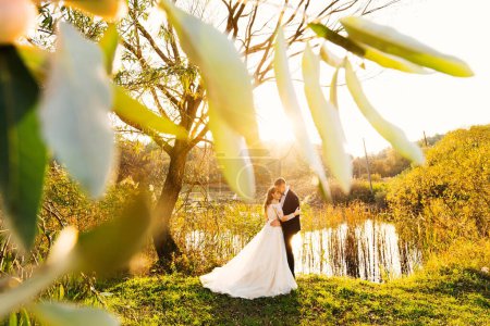 Photo for The bride in a wedding dress and the groom in a suit hug on the shore of the lake. evening lake. trees near the lake. - Royalty Free Image