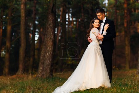 Foto de La novia en un vestido de novia y el novio en un traje se abrazan y cierran los ojos en el bosque - Imagen libre de derechos