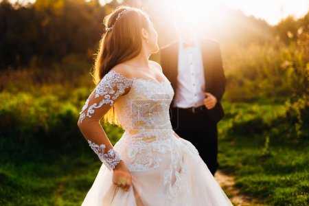 Téléchargez les photos : Mariée dans une belle robe avec une dentelle regardant par-dessus son épaule au marié. soleil du soir - en image libre de droit