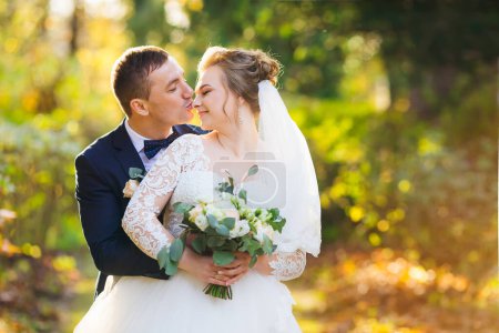 Téléchargez les photos : Le marié étreint la mariée et l'embrasse sur le nez. mariée a fermé les yeux et tient un bouquet de mariage. jeunes mariés dans le parc. - en image libre de droit