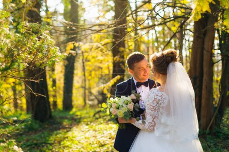Foto de La novia sostiene un ramo de bodas y el novio la abraza y sonríe. recién casados en el parque. - Imagen libre de derechos