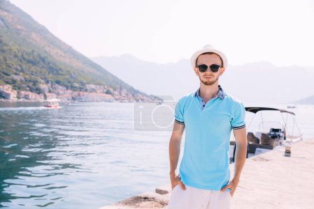 Foto de Hombre guapo en una camiseta azul se encuentra en el puerto deportivo del yate cerca del agua de mar. Vacaciones marítimas. Vacaciones de verano. Copiar espacio. - Imagen libre de derechos