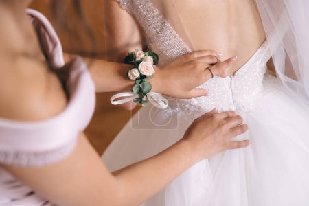 Photo for Bridesmaid helping bride fasten buttons on corset and getting her dress, preparing bride in morning for the wedding day. - Royalty Free Image