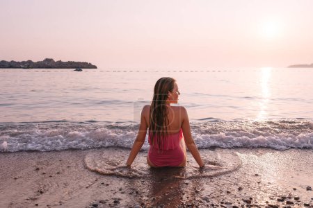 Téléchargez les photos : Jeune femme assise dans le sable regardant la mer au coucher du soleil - en image libre de droit