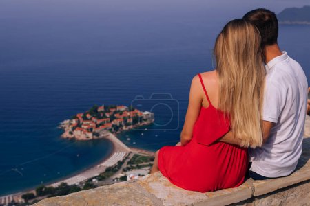 Foto de Rear view romantic young couple in love over sea shore above Sveti Stefan island in Budva, Montenegro. Travel. Vacation. Family. - Imagen libre de derechos