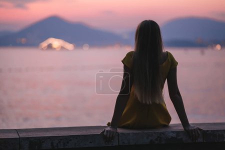 Foto de Una joven hermosa mujer en un vestido se sienta en la playa al atardecer y mira el sol poniente, chica solitaria sueña con el amor. - Imagen libre de derechos