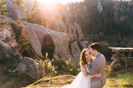 Téléchargez les photos : Jeune beau couple d'homme et de femme dans une robe de mariée et costume étreignant dans la nature près des hautes montagnes de pierre. - en image libre de droit