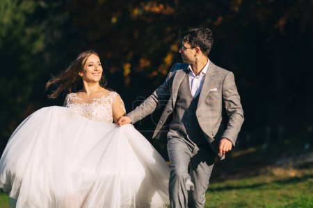 Photo for Newly married couple running and jumping in park while holding hands - Royalty Free Image