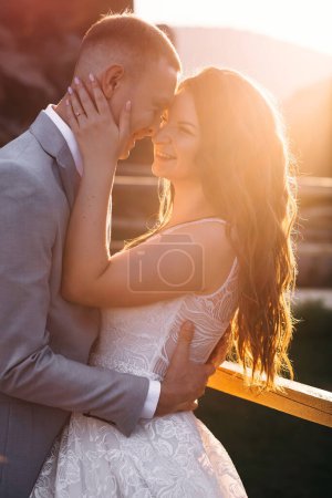 Foto de Preciosos recién casados se paran uno frente al otro, abrazándose en el fondo de la puesta de sol. Siluetas románticas. - Imagen libre de derechos