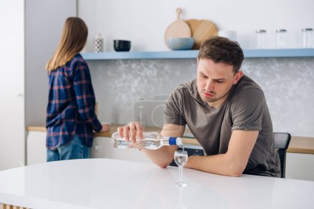 Téléchargez les photos : Disgruntled housewife standing behind and watching her alcoholic husband. A couple of a man and a woman have conflicts in the home kitchen due to male alcoholism. Relationship and marriage problems - en image libre de droit