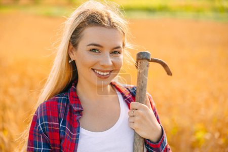 Foto de Girl gardener smiles at the camera, posing with a hoe while working in a wheat field. - Imagen libre de derechos