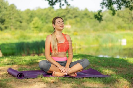 Foto de Primer plano retrato de la mujer sonriente en traje deportivo relajante meditación sensación zen-como en la alfombra de fitness en el parque público al aire libre. Vida activa saludable - Imagen libre de derechos