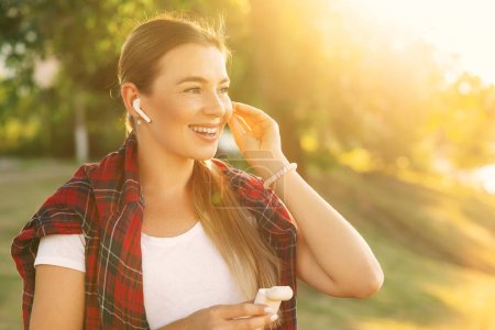 Foto de Mujer feliz sonriente de pie en el parque verde de la ciudad en verano escuchando música con auriculares y mirando hacia otro lado. Copiar espacio - Imagen libre de derechos