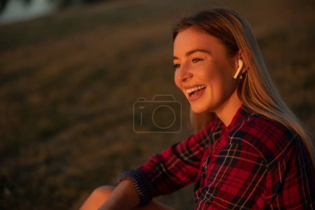 Foto de Foto retrato sonriente mujer feliz sentada en el parque verde de la ciudad en verano escuchando música en auriculares y admira la puesta de sol - Imagen libre de derechos