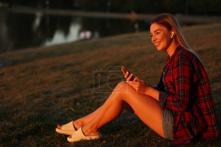 Téléchargez les photos : Souriante jeune femme blonde écoutant de la musique tout en étant assise sur l'herbe dans le parc près du lac et admire le coucher du soleil - en image libre de droit