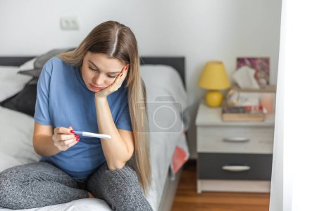 Téléchargez les photos : Upset blonde girl checking her recent pregnancy test, sitting on bed at home, copy space - en image libre de droit
