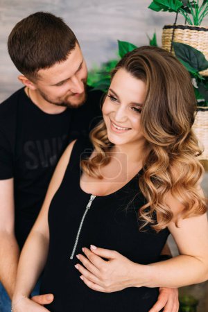 Photo for Handsome man and his beautiful pregnant wife hugging belly and smiling at home - Royalty Free Image