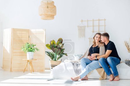 Téléchargez les photos : Une famille heureuse qui attend un enfant. Une femme enceinte est assise avec son mari dans un appartement confortable, serrant son estomac - en image libre de droit