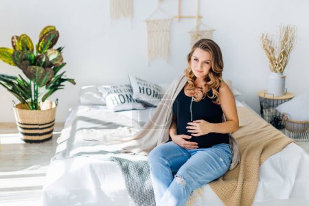 Foto de Feliz embarazada es sentado en el sofá en casa en el dormitorio abrazando su vientre - Imagen libre de derechos