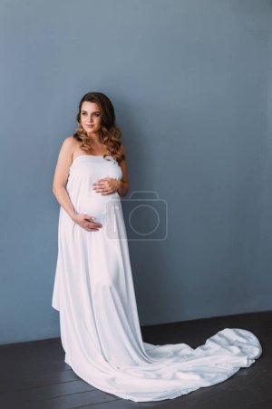 Foto de Una chica embarazada se cubre con una tela de seda blanca en un estudio sobre un fondo blanco - Imagen libre de derechos