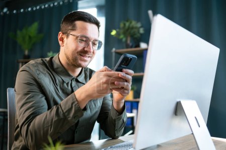 Foto de Foto de hombre adulto sonriente de 30 años en camisa verde usando computadora portátil y teléfono móvil, mientras está sentado en la mesa y trabajando en la oficina - Imagen libre de derechos