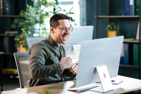 Téléchargez les photos : Beau jeune homme d'affaires assis à son bureau, célébrant le succès avec les bras levés en regardant son écran d'ordinateur - en image libre de droit