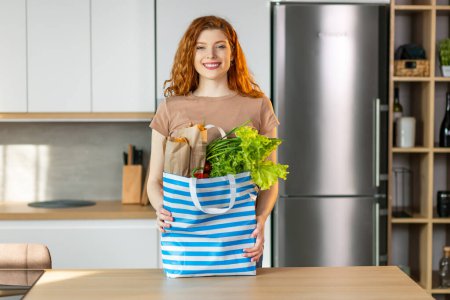 Téléchargez les photos : Belle jeune femme caucasienne tenir sac à provisions avec des légumes frais et des baguettes dans la cuisine moderne. - en image libre de droit